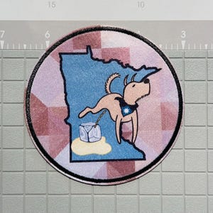 May include: A round patch featuring a cartoon dog in front of a blue outline of Minnesota. The dog wears a blue bandana with a star and is attached to a cube of ice. The background is a pink and white geometric pattern with a black border.
