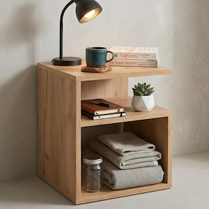 May include: A light brown wooden nightstand with two shelves. A black desk lamp, a teal mug, and books sit on top. The lower shelves hold a stack of folded blankets, a glass jar, a succulent in a white pot, and notebooks.