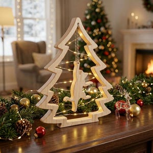 May include: A light-colored wooden cutout in the shape of a Christmas tree with a matching wooden cat figure hanging from the center. The frame is wrapped in thin, glowing string lights, creating a warm illumination around the cat silhouette.