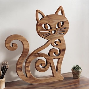 Puede incluir: Una silueta de madera marrón con forma de gato y veteado natural. La figura presenta recortes estilizados para los ojos, la nariz y los bigotes, junto con una cola larga y curvada y espirales decorativas integradas en el diseño del cuerpo.