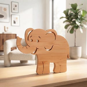 Op de afbeelding: Een lichtbruin houten olifantenfiguur met een gladde afwerking en een natuurlijke houtnerf. De olifant is afgebeeld in een staand profiel met de slurf omhoog, met uitgesneden details voor het oog, het oor en de mond, wat een eenvoudig silhouet creëert.
