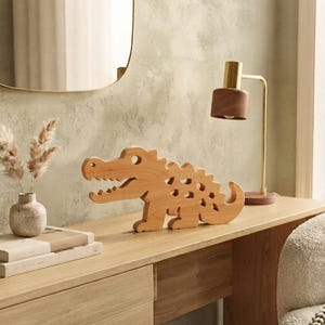 May include: A light brown wooden crocodile figurine with a cutout design along its back and tail. The figure features a wide open mouth with visible teeth and a rounded eye hole, crafted from smooth natural wood with a visible grain pattern.