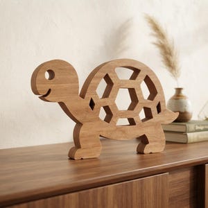 May include: A light brown wooden turtle cutout featuring a hollow geometric shell design. The turtle is depicted in profile, showing a smooth, natural wood grain finish, a rounded head with a circular eye cutout, and a stylized, open-work shell.