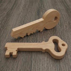 May include: Two wooden keys, one with a toothed edge and the other with a decorative handle. Both keys are made of light-coloured wood and lie on a wooden surface. The keys are likely decorative or for play.