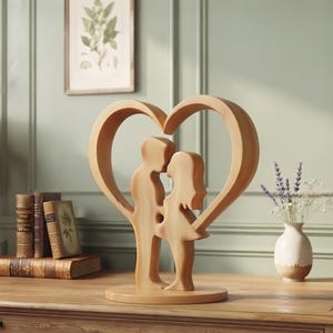 May include: A light brown wooden sculpture featuring two stylized figures standing face to face, enclosed within a large heart-shaped frame. The smooth, carved piece is mounted on a circular base, showcasing natural wood grain patterns throughout the design.