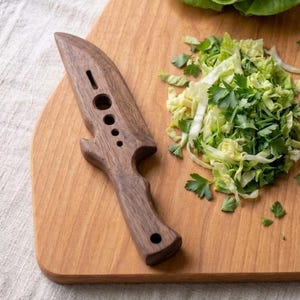 May include: A dark brown wooden kitchen tool shaped like a knife with a pointed tip, a curved handle, and a series of four circular holes of varying sizes along the center of the blade, resting on a light brown wooden cutting board.