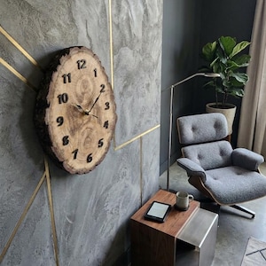Op de afbeelding: Een rustieke houten wandklok met een natuurlijke rand. De wijzerplaat is gemaakt van een dwarsdoorsnede van een boomstam, met zwarte cijfers en wijzers. De klok is gemonteerd op een grijze muur met gouden accenten, wat een moderne, natuurlijke uitstraling creëert.
