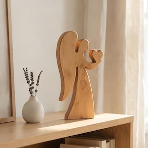 May include: A light brown wooden angel figurine with a smooth finish, featuring a stylized silhouette with a large wing and a small heart held in its hand. The figure is carved from wood with visible grain patterns and stands upright on a flat base.