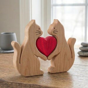 Könnte beinhalten: Zwei Holzkatzenfiguren, die ein rotes Herz halten. Die Katzen sind hellbraun mit einfachen Gesichtszügen und Schnurrhaaren. Das Herz ist leuchtend rot und bildet einen Kontrast zum Naturholz. Die Figuren stehen auf einer Holzoberfläche.