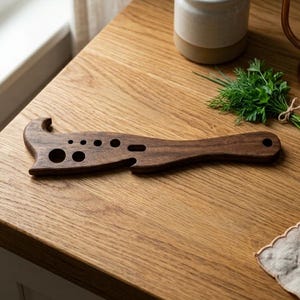 Puede incluir: Una herramienta de cocina de madera marrón oscuro con una forma única, con varios recortes circulares y un extremo en forma de gancho. La herramienta está diseñada para múltiples usos y descansa sobre una superficie de madera marrón claro.