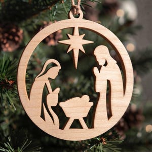 May include: A circular light brown wooden ornament featuring a laser-cut nativity scene. The design depicts two figures standing on either side of a central manger with a star positioned above them. A small loop at the top allows for hanging.