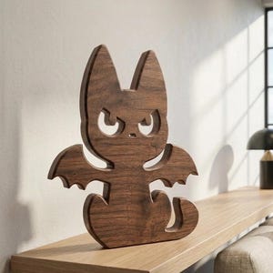 May include: A dark brown wooden cutout figure shaped like a stylised cat with bat-like wings. The figure features a smooth, polished wood grain finish, pointed ears, and cut-out eye shapes that create a distinct, sharp expression on the face.