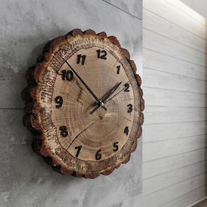 May include: A round wall clock crafted from a natural wood slice with visible bark edges. The clock face features a light brown wood grain pattern with dark brown, raised Arabic numerals from 1 to 12 and two thin black metal clock hands.