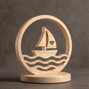 May include: A light-colored wooden decorative ornament featuring a circular frame enclosing a sailboat silhouette with a small heart cutout on the sail. The boat rests above three wavy lines representing water, all mounted on a circular wooden base.