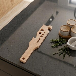 May include: A light-coloured wooden kitchen tool featuring a long handle with a circular hole at the end, a flat body with several circular cutouts of varying sizes, a rectangular slot, and a notched edge designed for gripping or hanging.