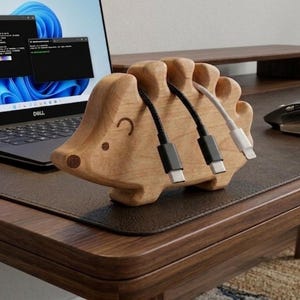 May include: A wooden hedgehog-shaped desk organizer with three slots holding black and white charging cables. The organizer is made of light-colored wood and has a simple, cartoonish design. A laptop is visible in the background.