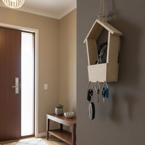 May include: A wooden key holder shaped like a house, hanging on a wall. The key holder has a small shelf and three hooks with keys attached. Sunglasses rest on the shelf. The key holder is made of light-colored wood.