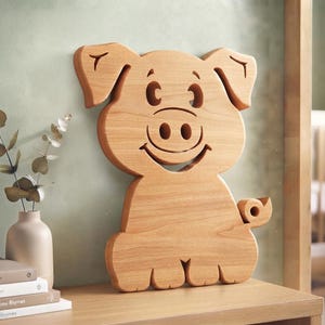 May include: A light brown wooden cutout shaped like a cartoon pig with large ears, a snout, and a curled tail. The pig features cut-out details for the eyes, nostrils, and a smiling mouth, showcasing a smooth, natural wood grain finish throughout.