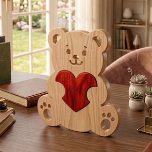 Puede incluir: Una figura de oso de madera de color claro con un diseño recortado y un acabado liso y pulido. El oso presenta una incrustación de madera en forma de corazón de color rojo intenso centrada en el pecho y recortes circulares para los ojos, la nariz y las patas.