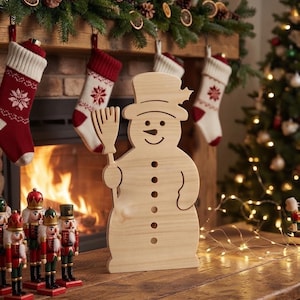 May include: A light-coloured wooden snowman cutout featuring a top hat, a broom held in one hand, and a series of circular cutouts down the centre representing buttons. The snowman has a simple engraved face with a curved smile and round eyes.
