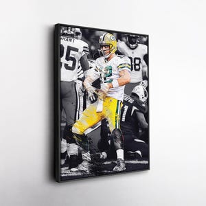 May include: A framed print featuring a football player in a green and yellow uniform, captured mid-play. The player is holding a football, with other players in the background. The image is in colour, set against a black and white backdrop.
