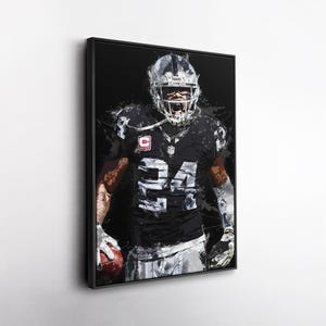 May include: A framed art print of an American football player in a black and silver uniform, number 24. The artwork has a splatter paint effect, with the player holding a football. The Raiders logo is visible on the helmet.