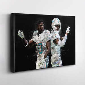 May include: Framed artwork featuring two football players in white and teal uniforms against a black background. The players are in action poses, with one raising his hand. The artwork has a painterly style.