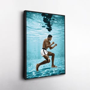 May include: Framed print of a man boxing underwater. The image features a man in white shorts with a black stripe and a black belt. The background is a light blue pool with water ripples.