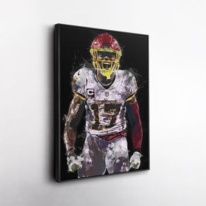 May include: Framed artwork of a football player in a Washington Commanders uniform, number 17. The player is wearing a red helmet and white jersey with gold accents. The background is black with white paint splatter effects.