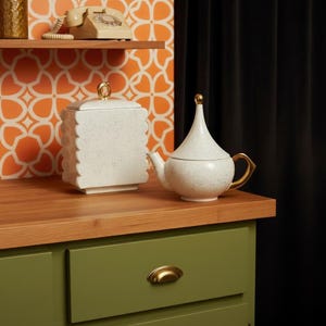 May include: A white speckled ceramic teapot with gold accents and a matching lidded container. The teapot has a curved spout and handle. The container has a scalloped edge and a gold handle. The items are on a wooden surface.