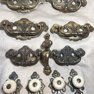 May include: A collection of antique brass drawer pulls and knobs. The drawer pulls have a decorative floral design and curved handles. The knobs have a white speckled ceramic center with a brass base. Ideal for vintage furniture restoration.