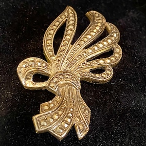 May include: A vintage gold-toned brooch in a bow design, encrusted with small, sparkling stones. The brooch has a detailed, textured surface and is set against a dark background. A classic accessory.
