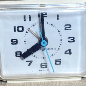 May include: A vintage GE Electric alarm clock with a white face and a cream-colored frame. The clock face features black numerals, black hands, and a blue second hand. The words "GE ELECTRIC" and "MADE IN U.S.A." are printed on the face.