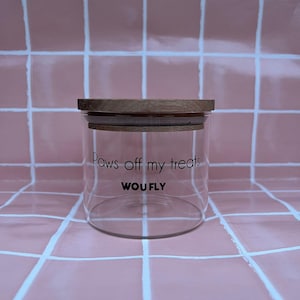 May include: A clear glass jar with a wooden lid. The jar has the text "Paws off my treats" and the brand name "WOUFLY" printed on the front. The jar is sitting on a pink tiled surface.