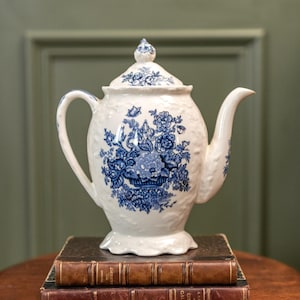 Puede incluir: Una tetera de cerámica blanca con un diseño floral azul, que incluye tapa y boquilla. La tetera está colocada encima de dos libros antiguos con cubiertas marrones, sobre una mesa de madera. La tetera mide aproximadamente 25 cm de alto.