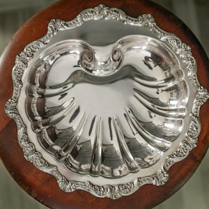 Puede incluir: Una fuente plateada ornamentada con un diseño en forma de concha. La fuente tiene un borde decorativo festoneado y se encuentra sobre una superficie de madera redonda de color marrón oscuro. La fuente refleja el entorno.