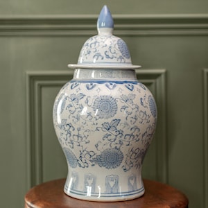 Puede incluir: Un jarrón de cerámica blanco y azul con tapa, con un diseño floral. El jarrón está sobre una mesa de madera. El diseño incluye motivos florales azules y bordes decorativos. Mide aproximadamente 30 cm de altura.