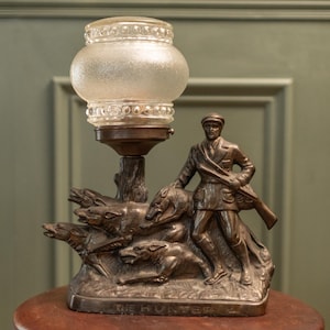 Op de afbeelding: Antieke bronzen lamp met een jager met honden, die een geweer vasthoudt. De lamp heeft een matglazen kap en staat op een houten tafel. De basis van de lamp luidt "THE HUNTER".