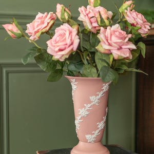 Könnte beinhalten: Eine rosa Vase mit weißen floralen Reliefs hält einen Strauß rosa Rosen und grüne Blätter. Die Vase steht auf einem dunklen Buch, mit einer grünen Wand im Hintergrund. Die Rosen sind in voller Blüte.