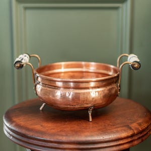 May include: A copper bowl with two handles and three small legs. The handles have white and black patterned ceramic ends. The bowl sits on a dark wooden stool against a green wall.
