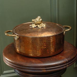 Op de afbeelding: Een koperen pot met een gehamerde afwerking staat op een houten kruk. De pot heeft een deksel met een decoratieve, goudkleurige vogelvormige handgreep en bijpassende handgrepen aan de zijkanten. De pot is rond en heeft een glanzend, reflecterend oppervlak.