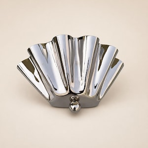 May include: A polished chrome metal shell-shaped dish with a reflective surface and a scalloped edge. The object features a small, spherical metal knob at the base, providing a stable point of contact. The design is symmetrical with deep vertical ridges.