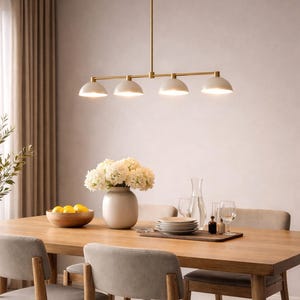 May include: A modern dining room setup featuring a wooden table and chairs. Above the table hangs a gold-toned linear chandelier with four white dome-shaped lights. A vase of white flowers and a wooden bowl of lemons adorn the table.