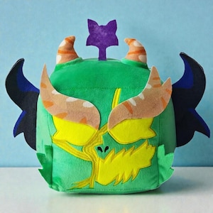 May include: A green plush monster toy with yellow facial features, orange and white horns, and black wings. A purple star-shaped detail sits on top. The soft fabric toy has a whimsical design and is suitable for children.