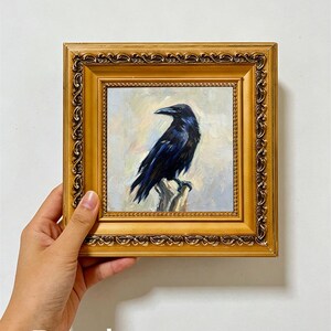 Può includere: Un dipinto a olio raffigurante un corvo nero appollaiato su un ceppo, inserito in una cornice quadrata ornata di colore oro metallizzato. L'uccello è dipinto con toni blu scuro e nero su uno sfondo chiaro, strutturato e neutro.