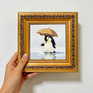 Puede incluir: Una pintura al óleo cuadrada en un marco dorado decorativo que muestra un pingüino sosteniendo un paraguas marrón. El pingüino tiene plumas blancas y negras con una mancha amarilla en el cuello, sobre una superficie de hielo azul claro y fondo gris.