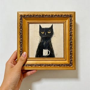May include: A square painting in an ornate gold frame depicting a black cat with yellow eyes holding a white mug. The cat has a stern expression and is rendered in a textured, painterly style against a neutral off-white background.