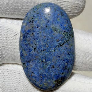 Pode incluir: Uma pedra preciosa polida de formato oval com uma cor base azul profunda e texturas manchadas de inclusões minerais pretas, verde-escuras e cinza-claras por toda a superfície, exibindo um acabamento liso e reflexivo em toda a pedra.