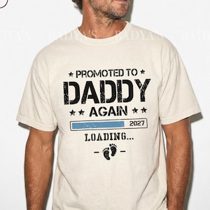 May include: Ivory-colored t-shirt with the text "PROMOTED TO DADDY AGAIN" in black, along with a loading bar and the year 2027. Below the loading bar are two baby footprints. A perfect gift for a new dad.