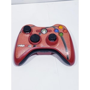 Peut inclure: Manette Xbox 360 rouge et noire avec un motif de ballon de basket. La manette comporte des joysticks analogiques noirs, des boutons et un pavé directionnel. Le texte "NBA 2K13" est visible.